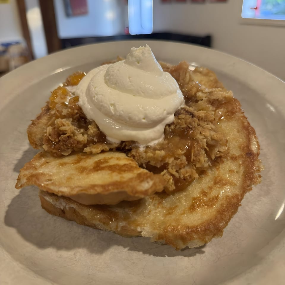 A golden-brown French toast topped with apple crumble, caramel sauce, and a dollop of whipped cream.