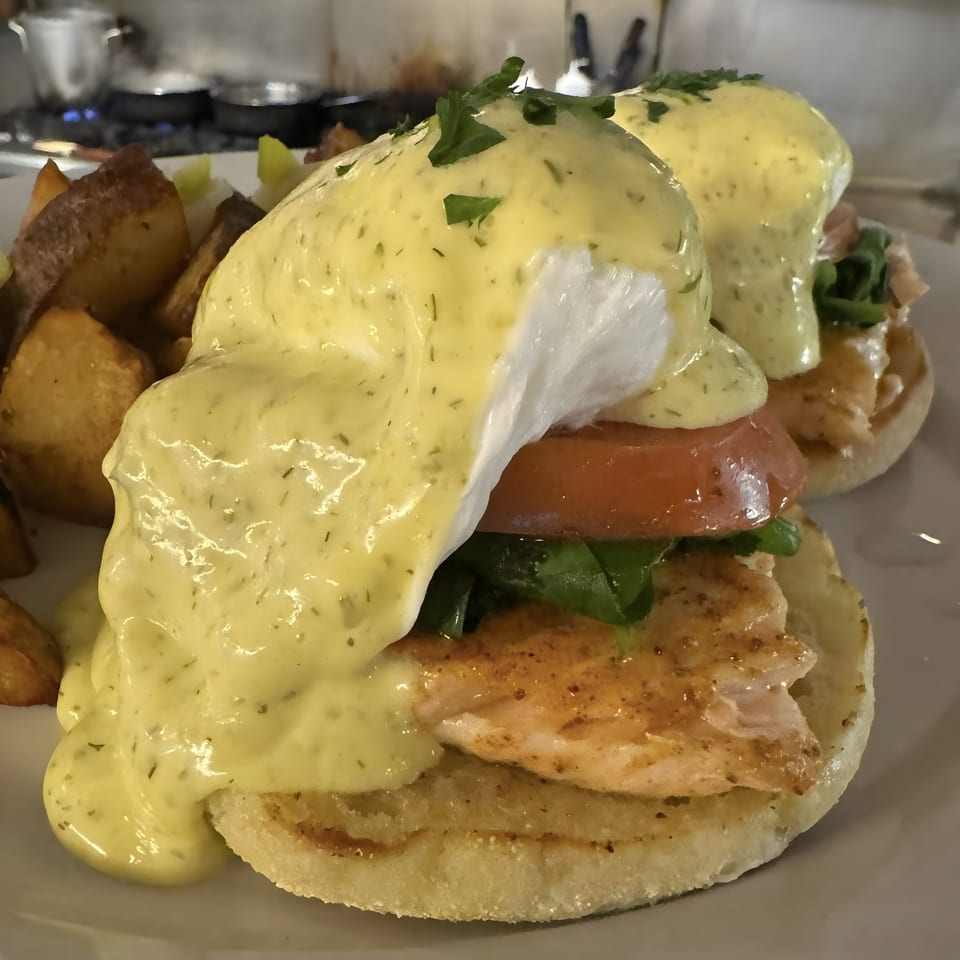 Two servings of Eggs Benedict with salmon, tomato, and spinach, topped with hollandaise sauce and a side of roasted potatoes.