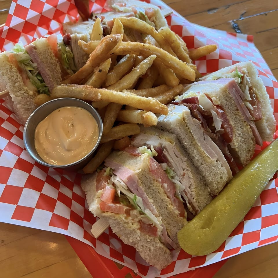 Acme Cafe's club sandwich with fries, a side of sauce, and a pickle spear in a red checkered basket.